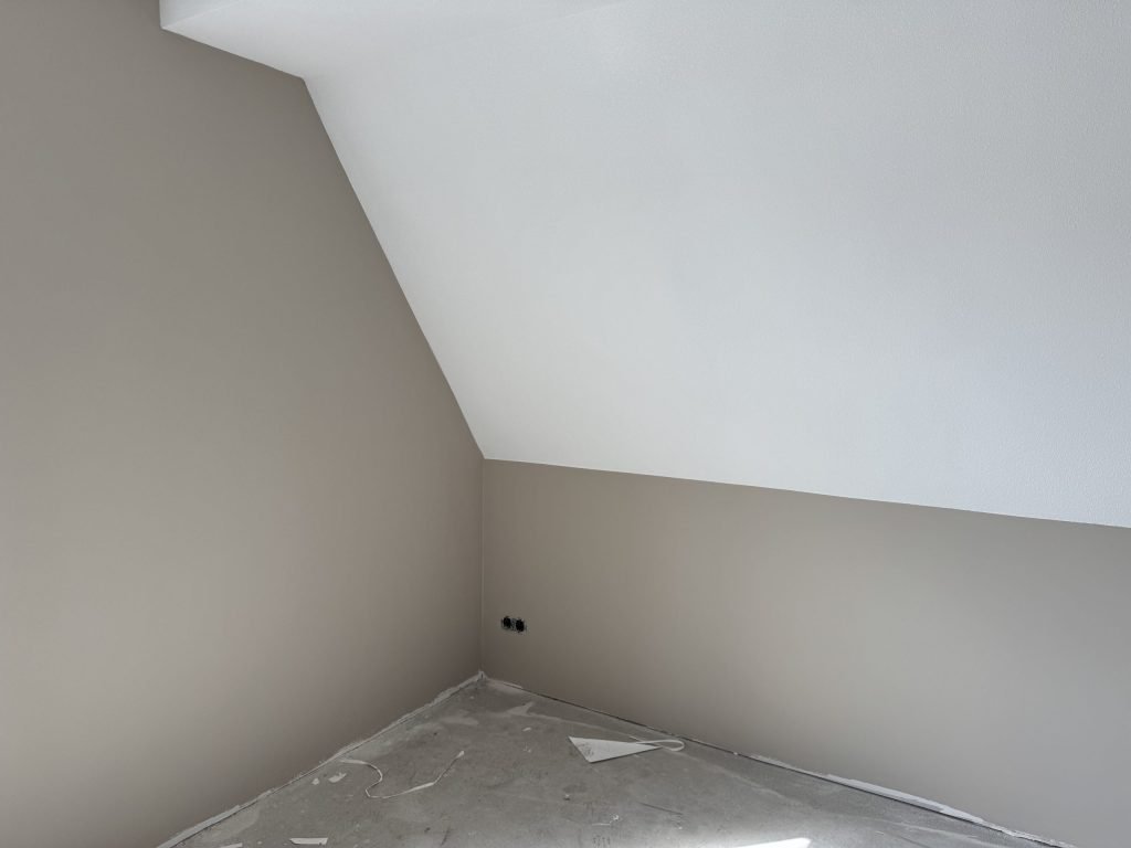 Zolderkamer met een witte schuine kant en een grijs gesausde wand, vloer nog afgedekt