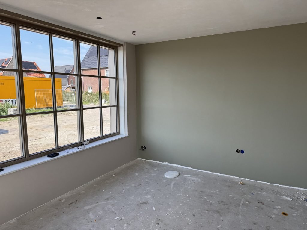 Nieuwbouwkamer met een egale grijsgroene wand naast een groot raam met zwarte kozijnen