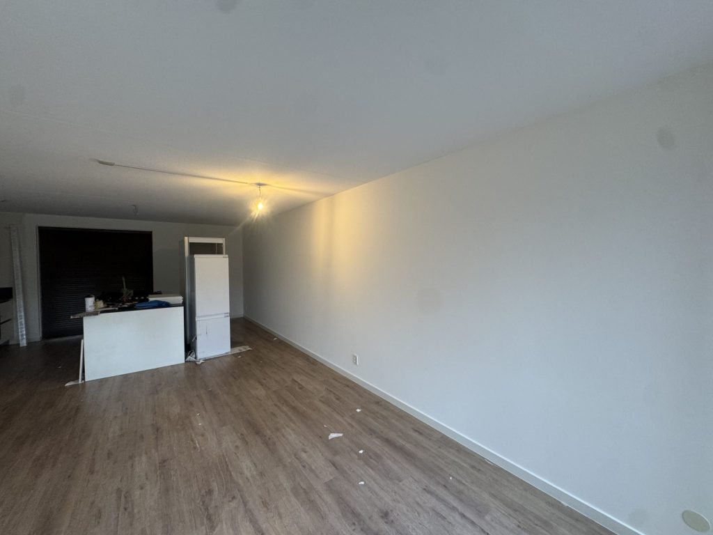 Woonkamer met een laminaatvloer, strakke witte wanden en een keukenblok achterin