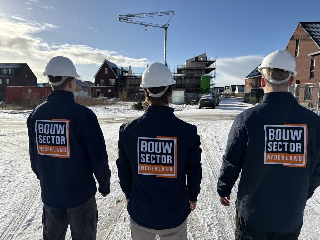 Drie vakmensen van Bouwsector Nederland met witte helm en logo op de rug bij een nieuwbouwwijk in de sneeuw